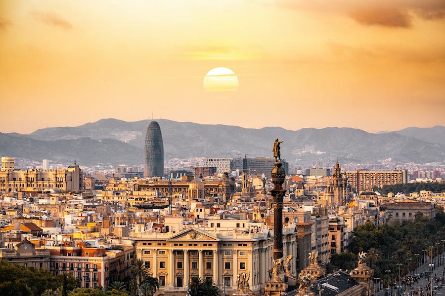 Entdecken Sie die atemberaubende Skyline von Barcelona bei Sonnenuntergang, mit Blick auf markante Sehenswürdigkeiten wie den Torre Glòries und die historische Columbus-Säule, eingebettet in die malerische Landschaft der umliegenden Hügel.